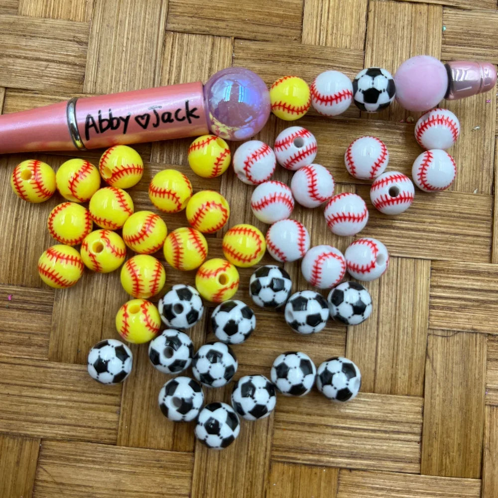 10MM acrylic sports beads in yellow softball, white baseball, and black and white soccer ball patterns arranged on a wicker surface next to a beadable pen.