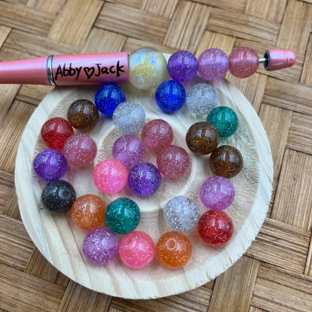 10MM round glitter acrylic beads in assorted colors including pink, purple, blue, teal, red, orange, brown, black, and iridescent, displayed on a wooden tray next to a pink beadable pen labeled Abby Jack.