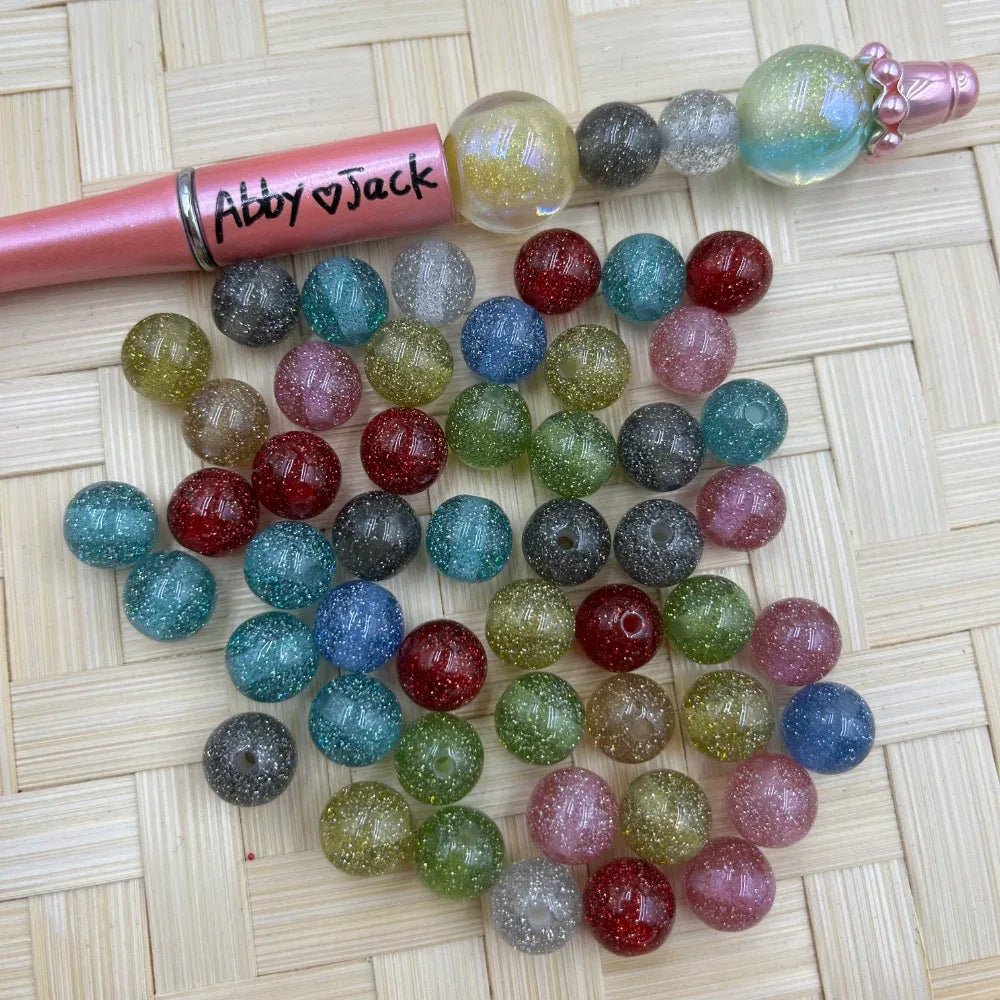 10MM mixed color glitter sparkle acrylic round beads displayed on woven surface next to a beadable pen, showing assorted translucent shimmer colors including teal, red, pink, green, gray, and blue.