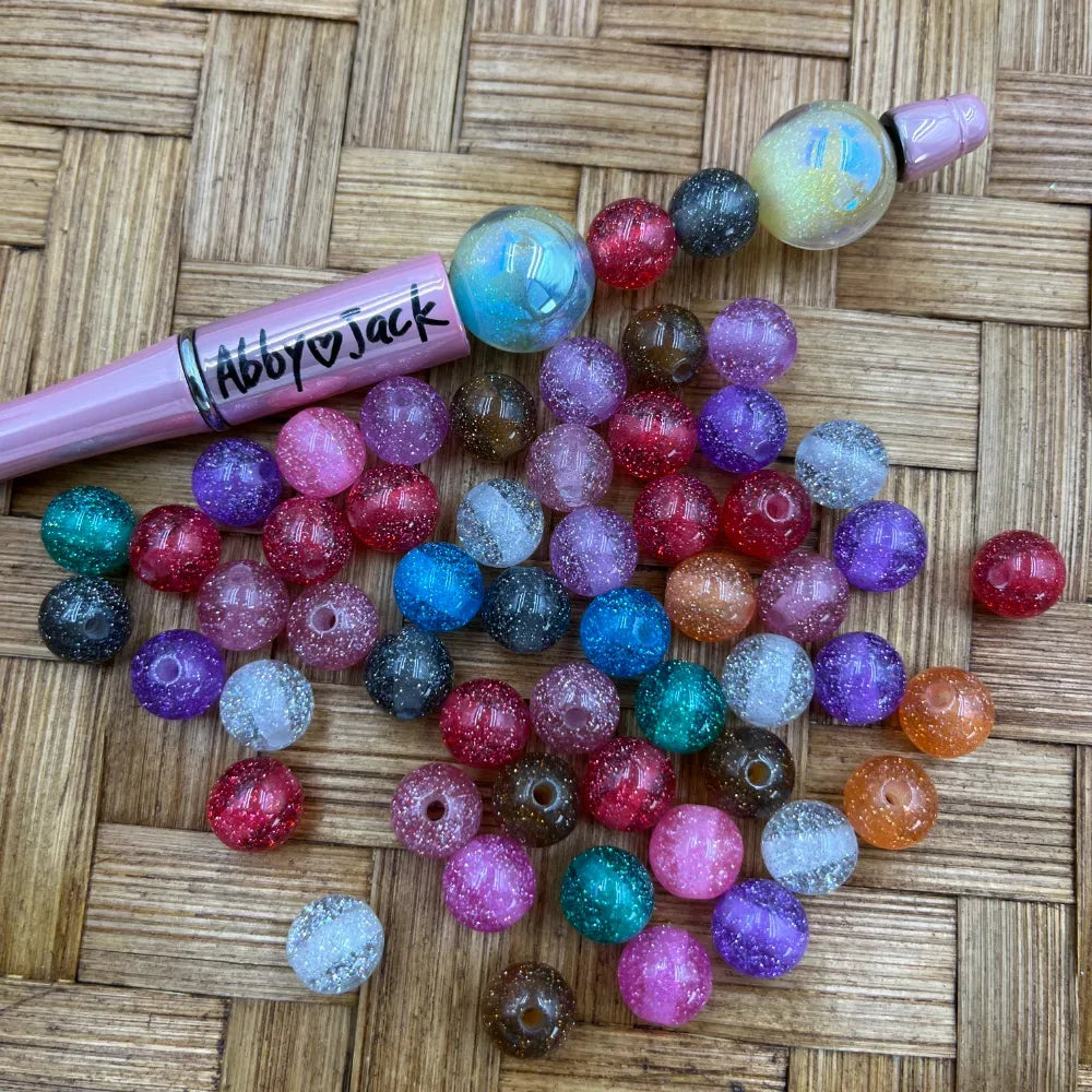 10MM multicolor glitter stardust acrylic round beads scattered on woven surface next to a pink beadable pen, showing assorted colors including red, purple, blue, teal, pink, orange, and clear with sparkle finish.