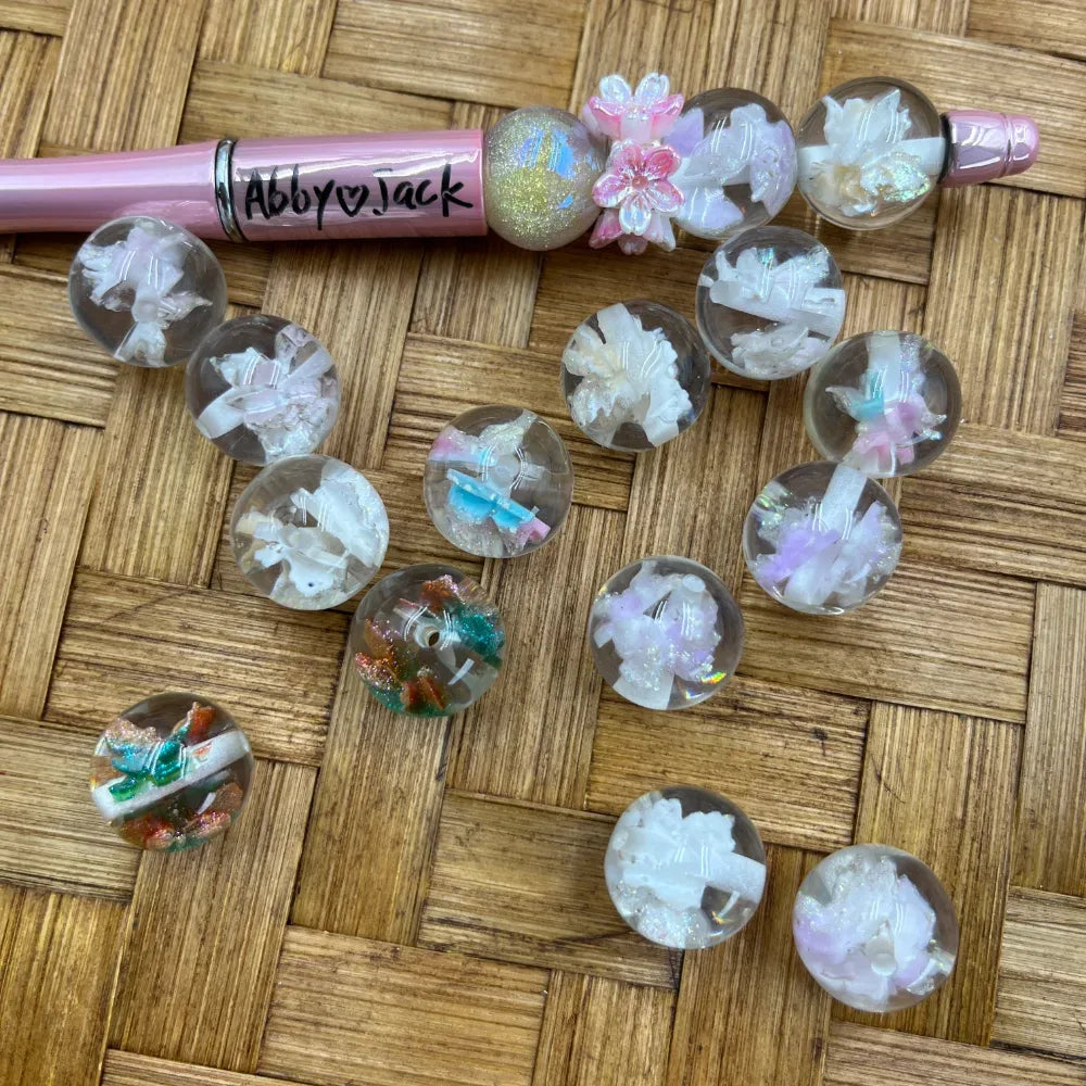 16MM clear acrylic round beads with encased white iridescent floral and crystal chip inclusions, displayed with a pink beadable pen on wicker surface