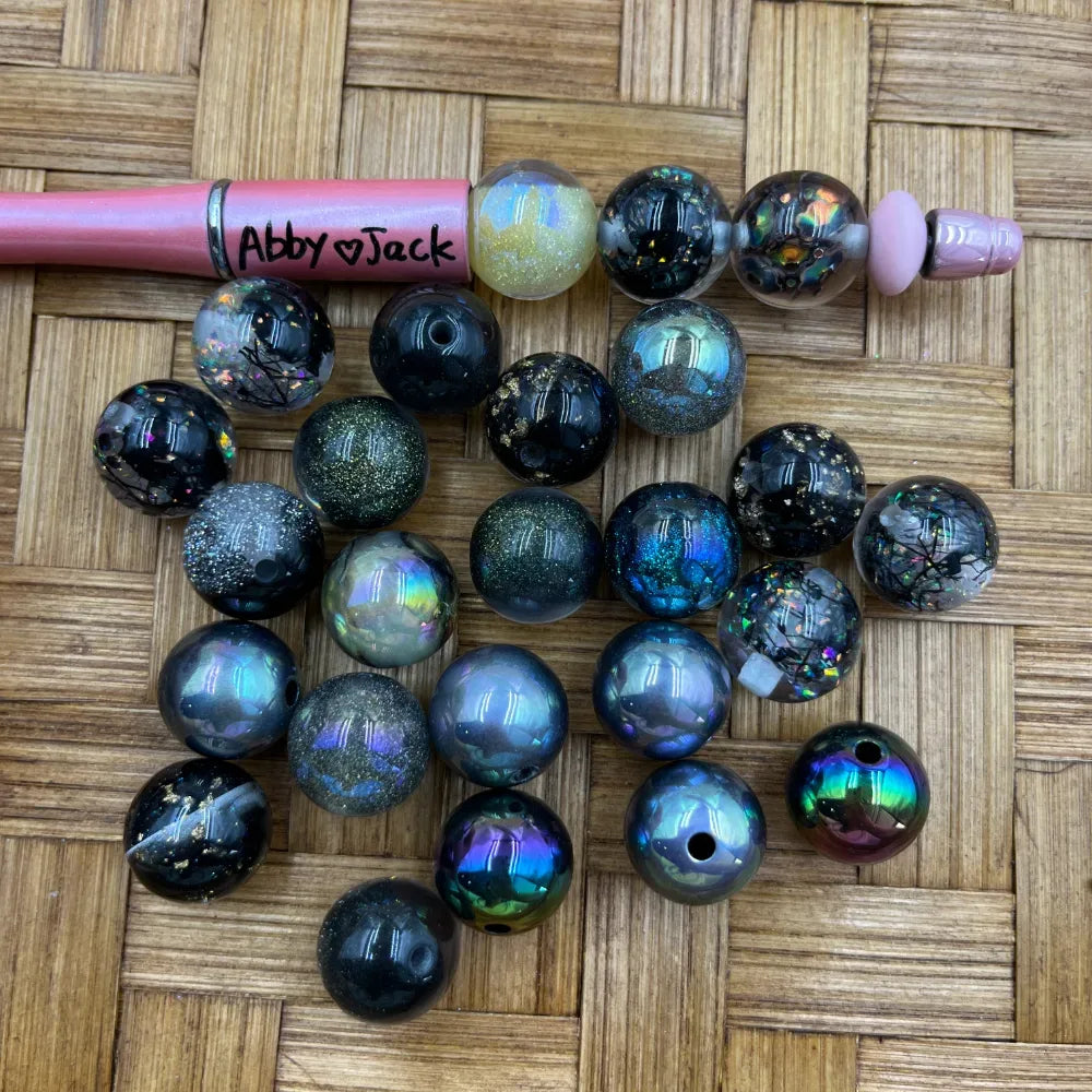16MM galaxy iridescent glitter acrylic beads in dark black, blue, green, and holographic finishes displayed on a woven surface next to a beadable pen for size reference.