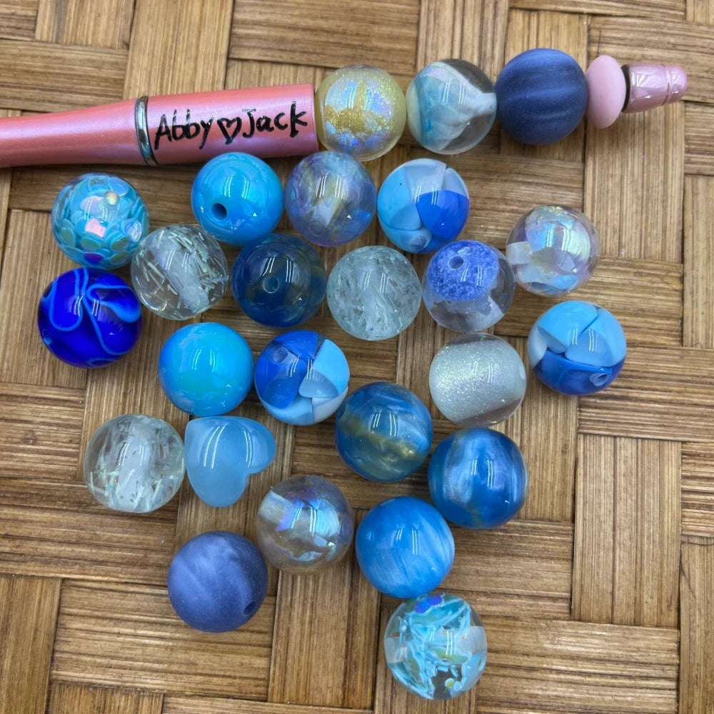 16MM blue ocean mix acrylic beads in assorted finishes including iridescent, glitter, marble, chunky, and crackle styles arranged on a woven surface next to a beadable pen