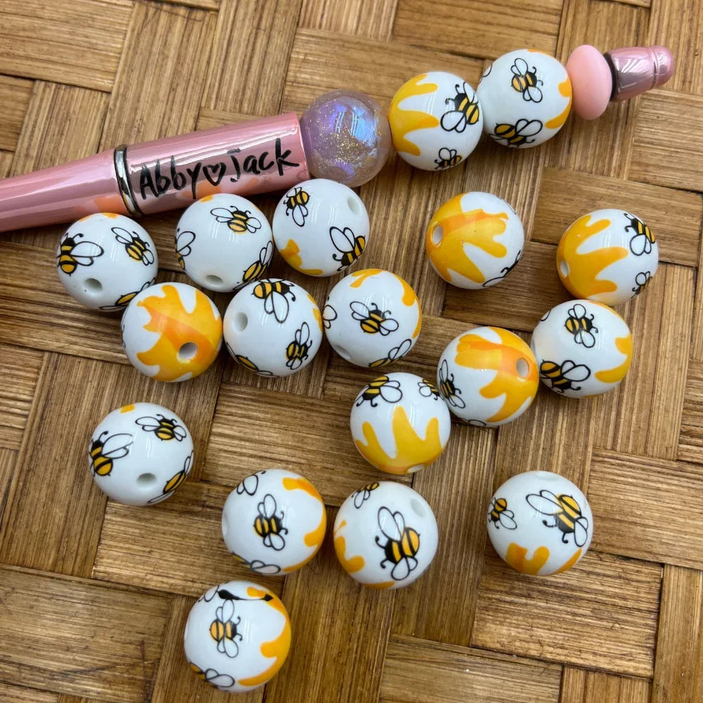16MM white acrylic round beads printed with yellow bumble bees and honeycomb pattern, shown scattered on wicker surface next to a beadable pen.