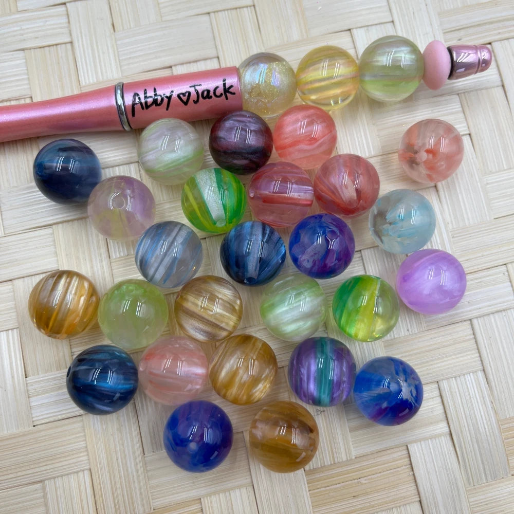 25 assorted 16mm multicolor swirl acrylic beads in mixed colors including blue, purple, green, pink, gold, and clear iridescent finishes, displayed on a woven surface next to a pink beadable pen.