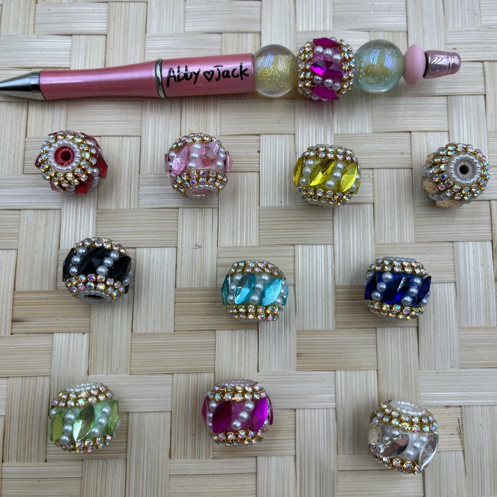 Assorted multicolor rhinestone and faux pearl acrylic fancy beads shown beside a pink beadable pen, including pink, red, yellow, blue, black, green, clear, and white designs.