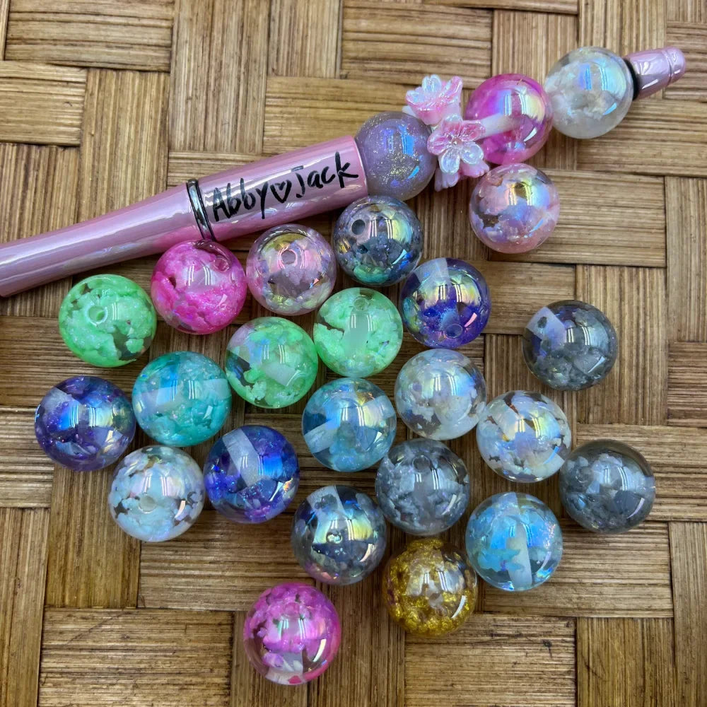 16MM aurora crystal crush acrylic beads in assorted colors including pink, green, blue, gray, and gold with iridescent shimmer effect, displayed with a pink beadable pen on woven bamboo surface.