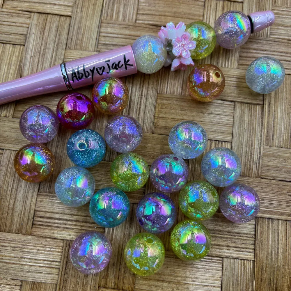 16MM aurora galaxy glitter acrylic beads in iridescent rainbow shimmer colors including teal, pink, purple, gold, and clear, displayed next to a beadable pen on a wicker surface.