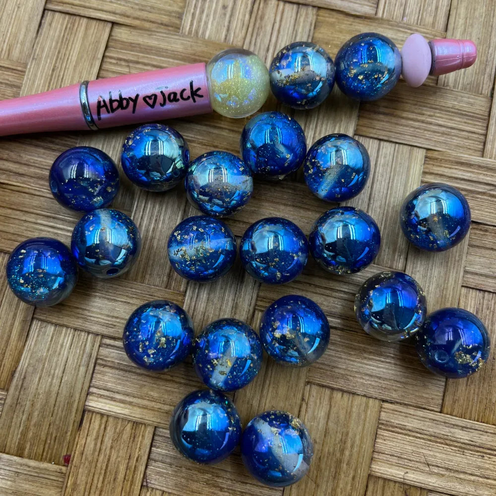 16MM deep blue galaxy acrylic beads with gold fleck foil inclusions scattered on a woven surface next to a pink beadable pen labeled Abby heart Jack