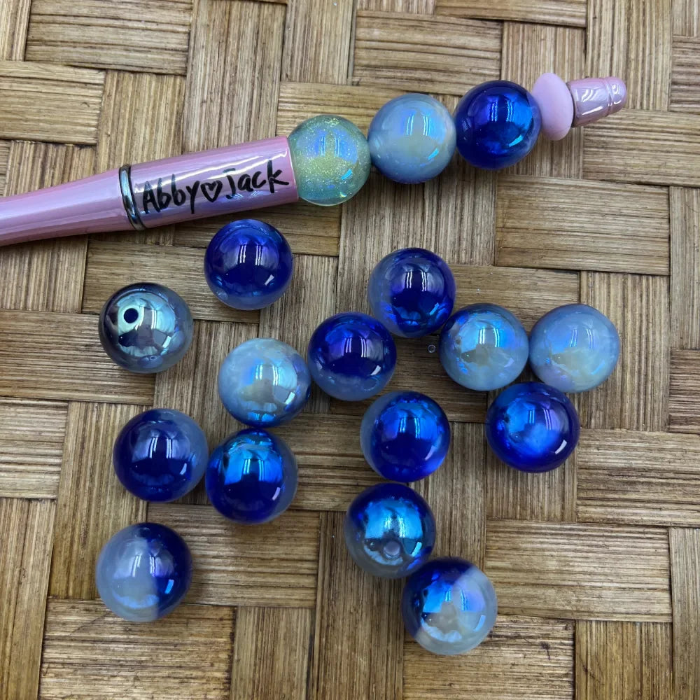 16MM cobalt blue and opal iridescent aurora acrylic beads scattered on wicker surface next to a pink Abby Jack beadable pen with beads threaded on it