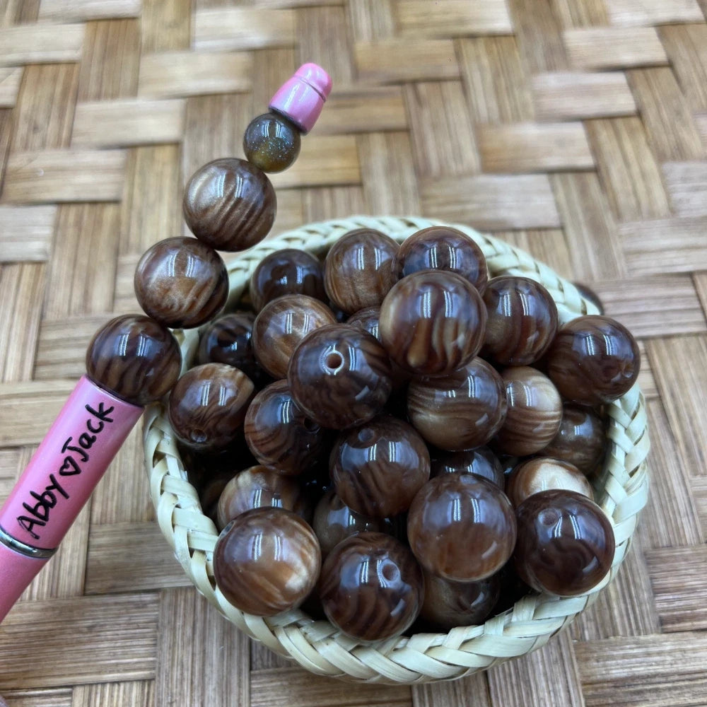 16MM brown wood grain swirl acrylic beads in a small wicker basket with a beadable pen, shown on a woven bamboo background.