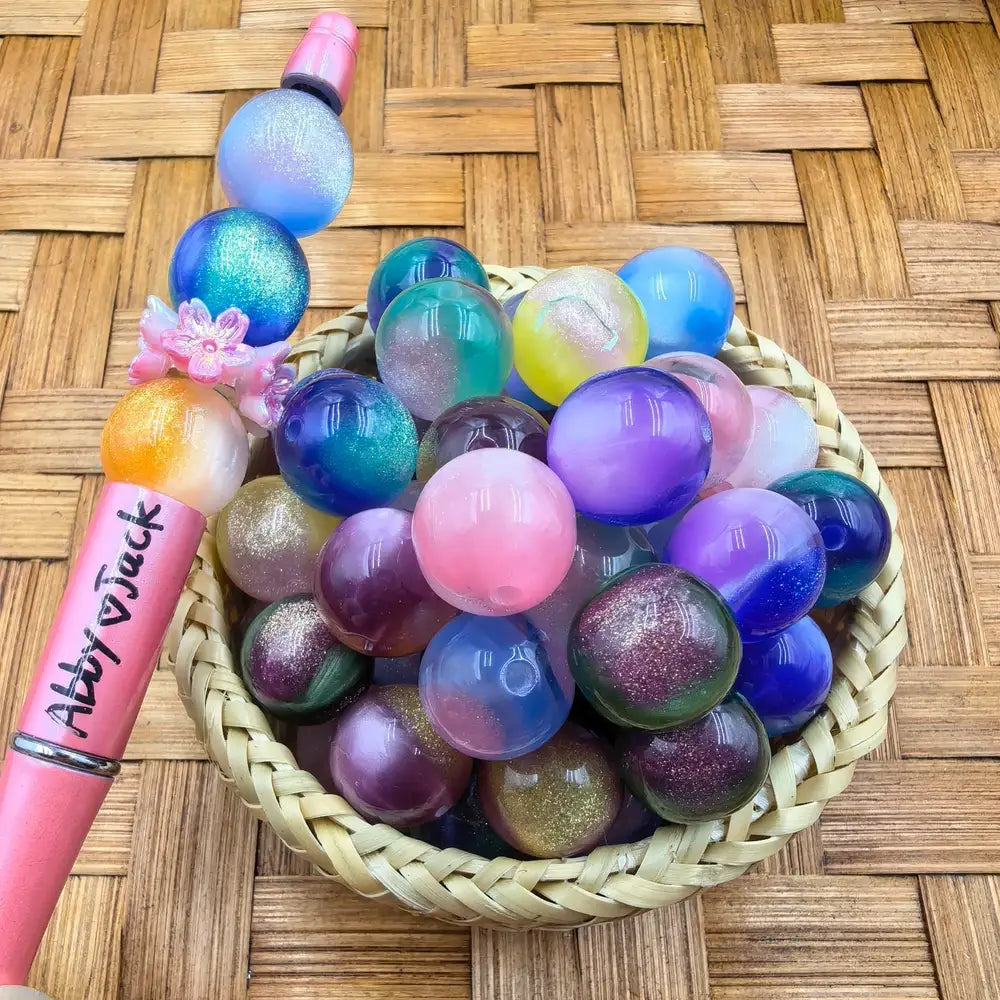 16MM galaxy glitter acrylic beads in a wicker basket displaying iridescent multicolor shimmer tones including purple, blue, pink, teal, gold, and green, shown next to a pink beadable pen.