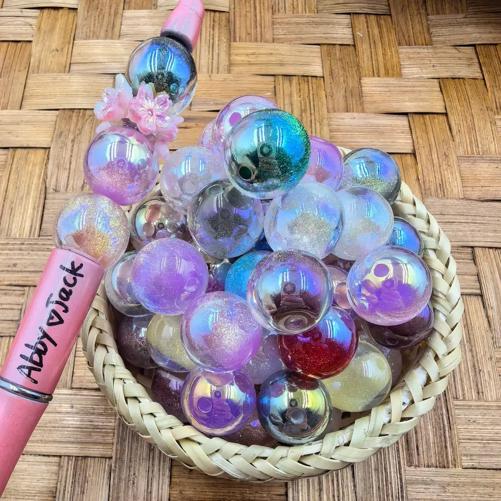16MM iridescent aurora glitter acrylic beads in holographic purple, blue, and clear finishes displayed in a wicker basket next to a pink beadable pen, perfect for DIY jewelry making and beadable pens.