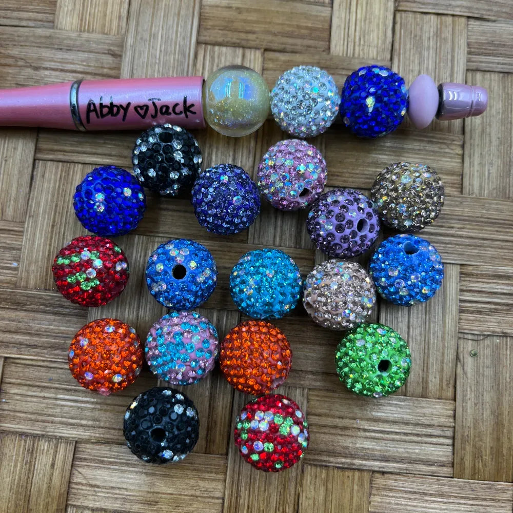 16MM mixed color rhinestone beads in assorted colors including blue, red, orange, green, black, pink, teal, and purple with glittery crystal rhinestone finish, displayed on a woven surface next to a beadable pen for size reference.
