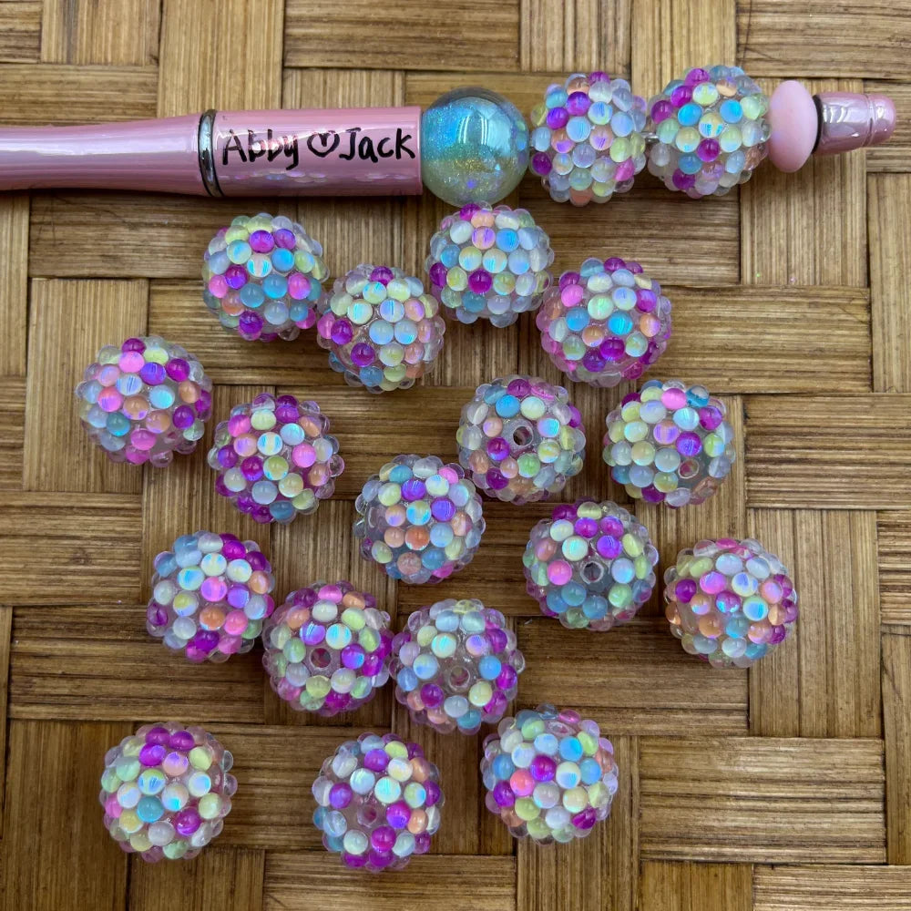 16MM rainbow AB iridescent cluster bubble fancy beads in pink, blue, yellow, and purple multicolor, shown loose and on a pink beadable pen on a woven bamboo surface.
