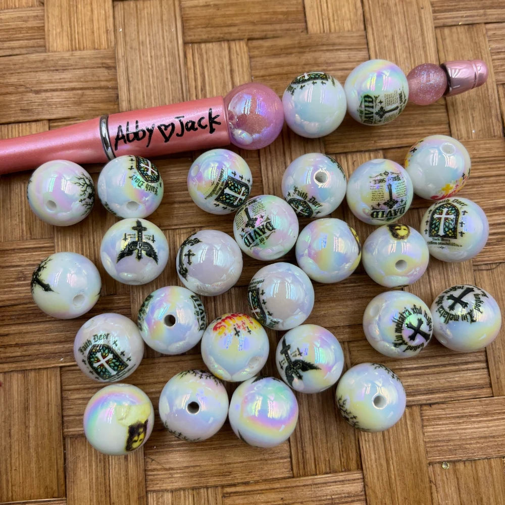 Iridescent AB finish white acrylic beads printed with Christian cross, Faith, and David religious designs, shown with a pink beadable pen on a wicker surface.