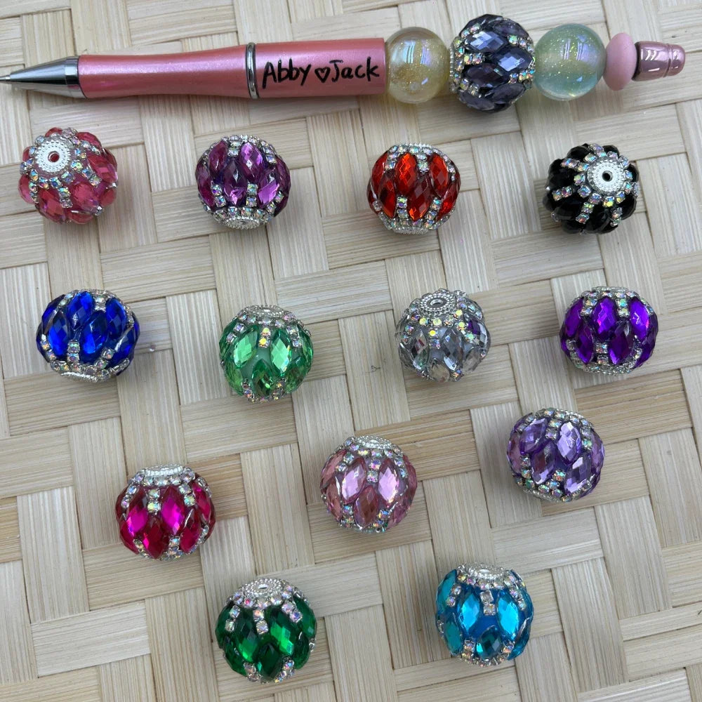 Assorted 20mm round faceted rhinestone acrylic fancy beads in pink, purple, red, black, blue, green, gray, and aqua beside a beadable pen