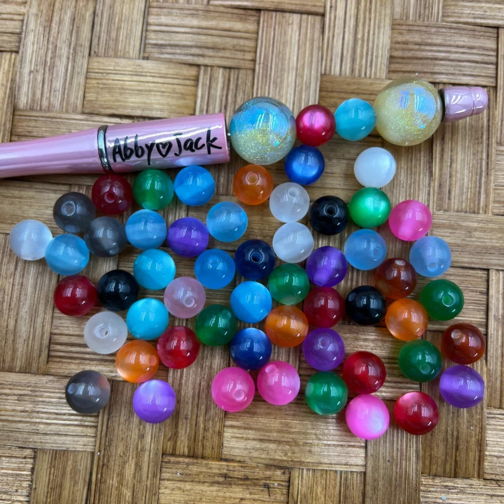 20MM mixed color cats eye acrylic beads in assorted colors including blue, red, green, purple, orange, pink, black, and white displayed on a wicker surface next to a pink beadable pen branded Abby Jack.
