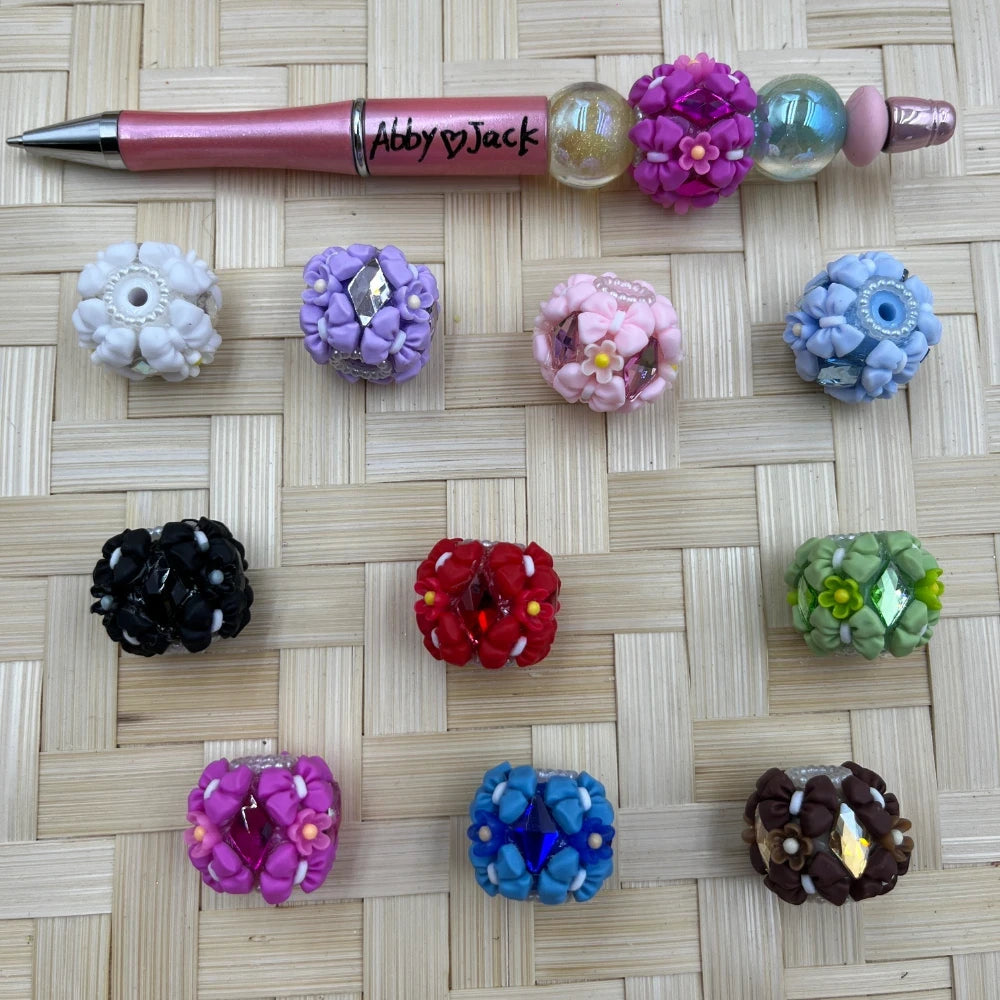 Assorted floral rhinestone clay fancy beads in white, lavender, pink, blue, black, red, green, fuchsia, royal blue, and brown shown with a decorated beadable pen.