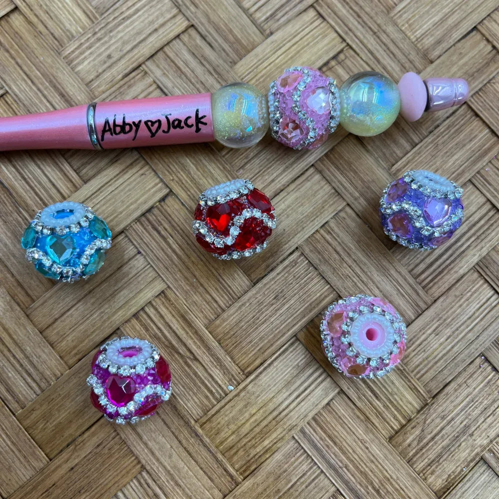 Assorted rhinestone bling crystal fancy beads in red, teal blue, pink, purple, and hot pink with silver rhinestone accents, displayed with a pink beadable pen labeled Abby and Jack on a wicker surface.