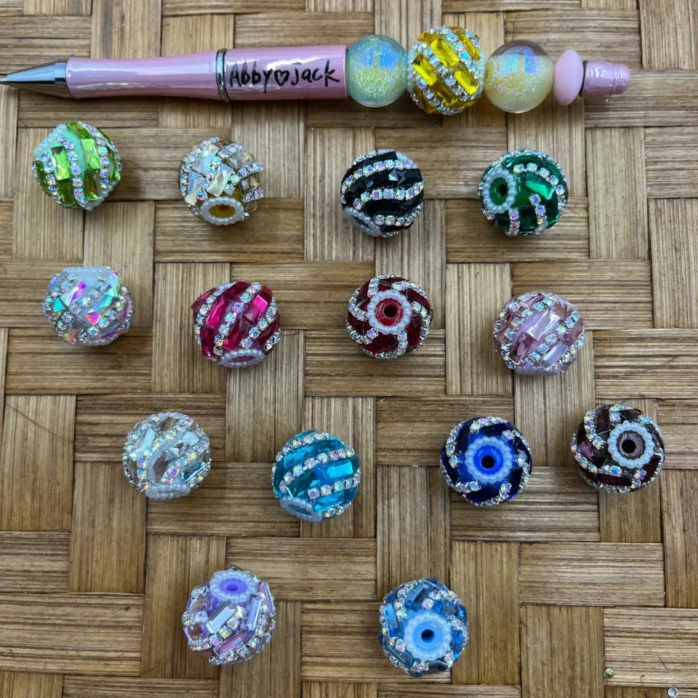 Assorted rhinestone crystal fancy beads in multiple colors including red, green, teal, blue evil eye, pink, black, and clear AB iridescent, displayed on wicker surface next to a beadable pen.