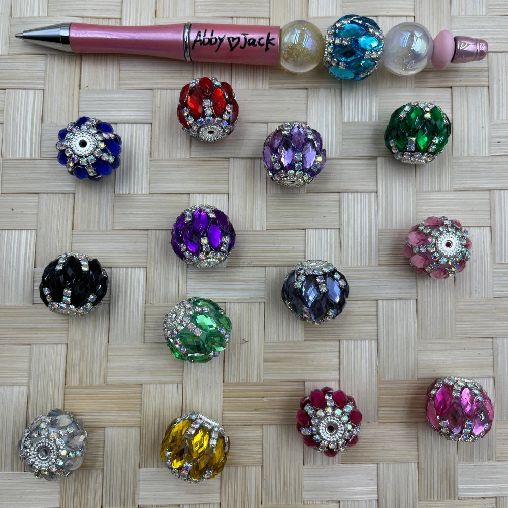 Assorted rhinestone crystal fancy beads in multiple colors including blue, red, purple, green, black, pink, yellow, and teal displayed on a woven surface next to a beadable pen.