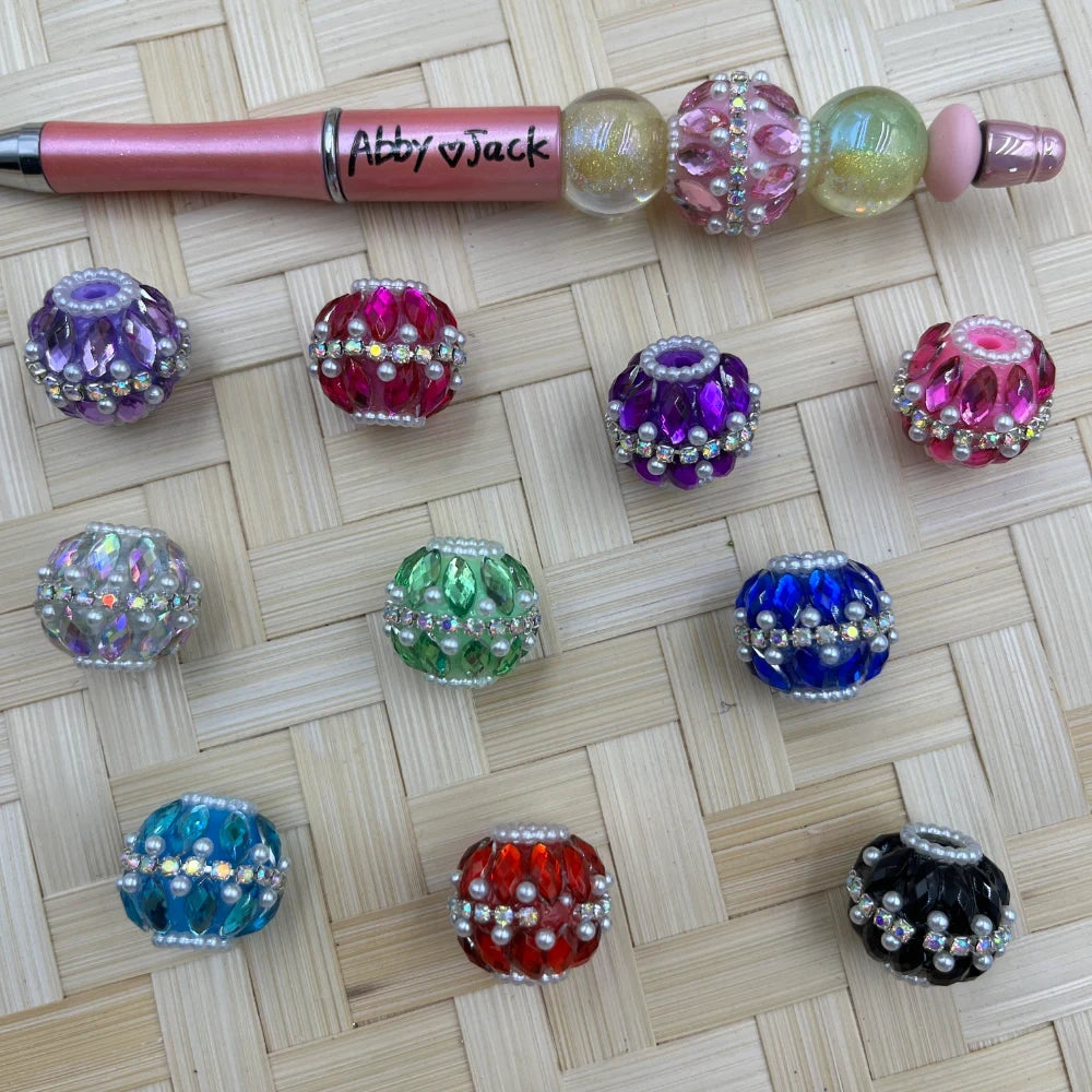 Assorted rhinestone and pearl-trimmed crystal fancy beads in purple, red, pink, green, blue, teal, orange, and black displayed on a wicker surface next to a decorative beaded pen labeled Abby and Jack.