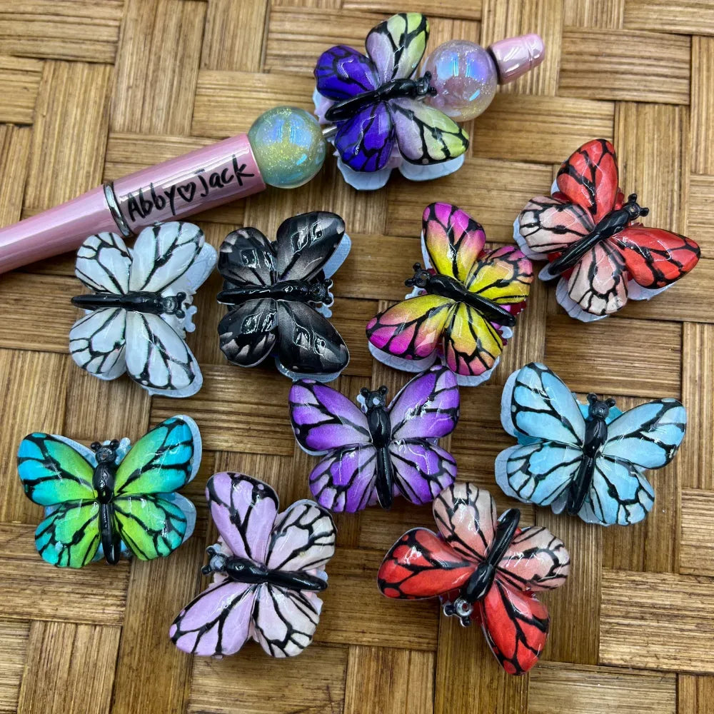 Colorful 3D butterfly fancy beads in multiple colors including purple, black, white, teal, pink, red, and blue displayed on a woven surface next to a beadable pen, perfect for DIY crafts and jewelry making.