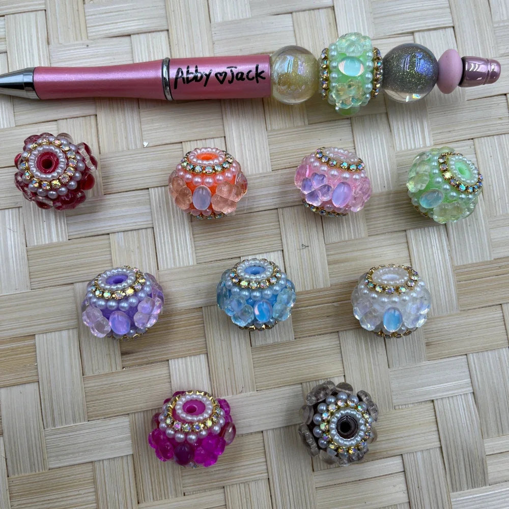 Assorted bubble-style glass fancy beads with pearl and rhinestone accents in multiple pastel and jewel tones displayed beside a pink beadable pen.