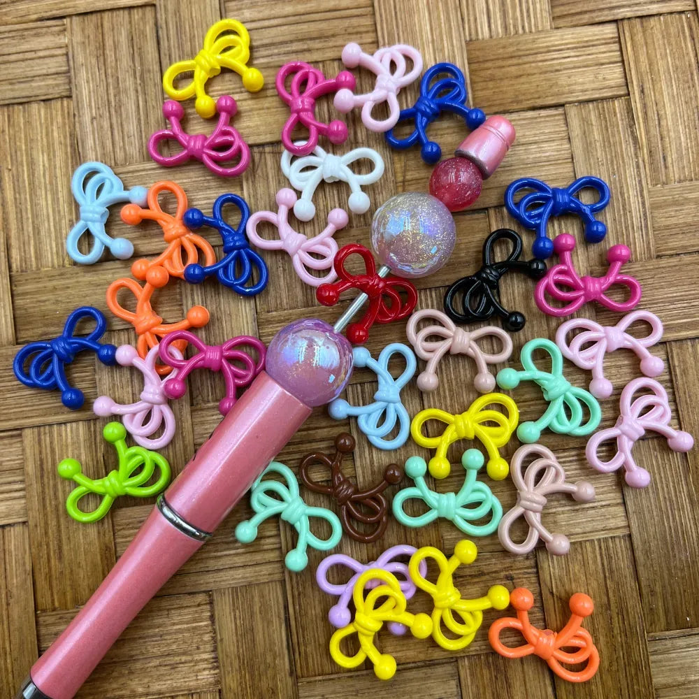Assorted colorful bow tie shaped acrylic beads in multiple colors including pink, blue, yellow, green, orange, red, black, white, and more, displayed on a woven bamboo surface next to a beadable pen with a glitter ball top.