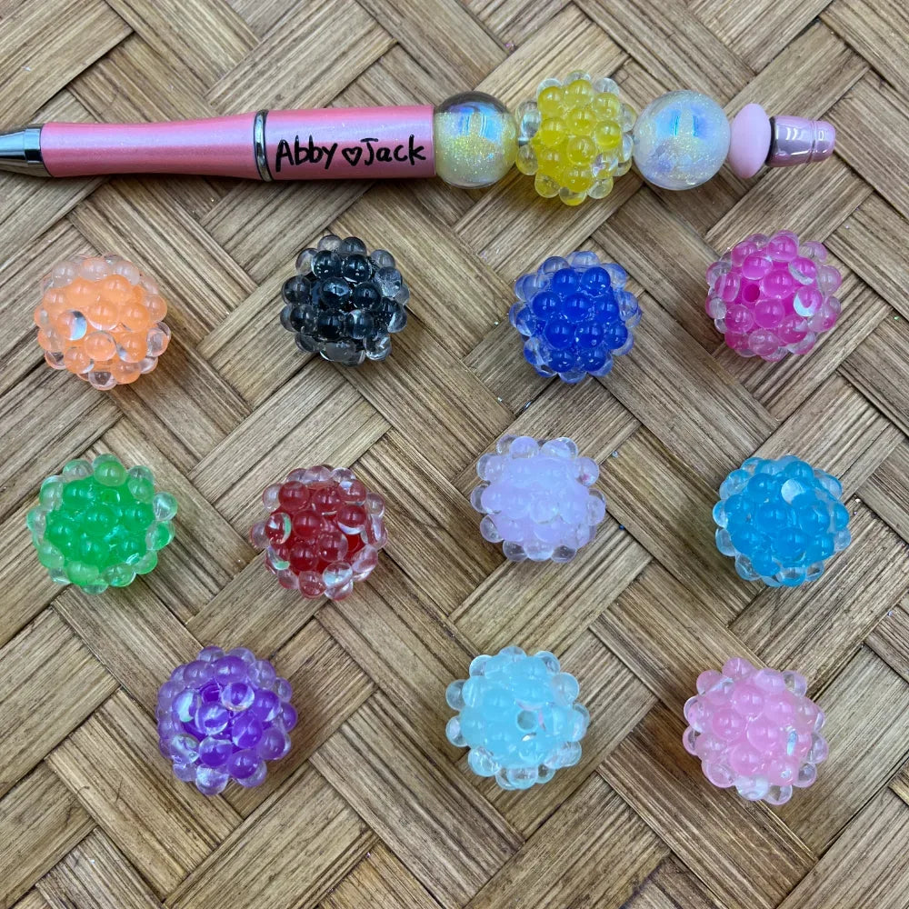 Assorted bubble cluster orbeez-style acrylic fancy beads in 11 colors including orange, black, blue, pink, green, red, white, light blue, purple, clear, and light pink, displayed on a woven rattan surface next to a pink beadable pen labeled Abby and Jack.