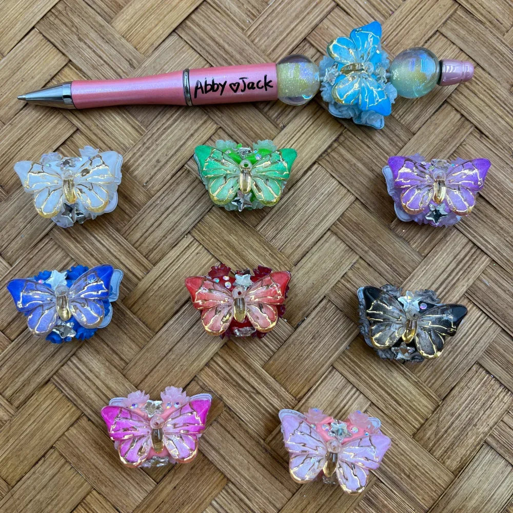 Assorted colorful butterfly shaped fancy acrylic beads with gold vein detailing in blue, green, purple, red, pink, black and white, displayed on woven bamboo surface next to a pink beadable pen labeled Abby and Jack.