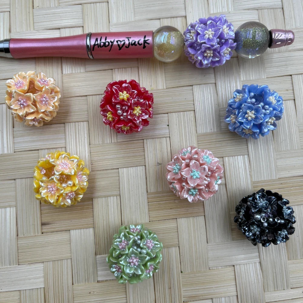 Assorted chunky 3D flower resin fancy beads in purple, peach, red, blue, yellow, pink, green, and black beside a pink beadable pen