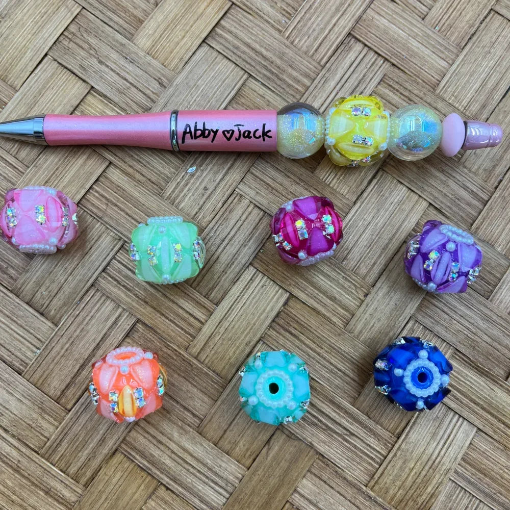 Colorful chunky rhinestone fancy beads in pink, green, magenta, purple, orange, teal, and blue with crystal embellishments, displayed next to a custom beaded pen labeled Abby and Jack on a wicker surface.