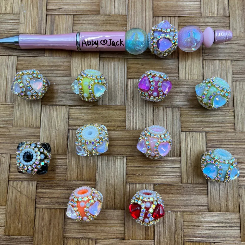 Assorted rhinestone-encrusted chunky fancy beads in multiple colors including pink, purple, green, red, black, and white opal with gold rhinestone borders, displayed on a woven bamboo surface next to a pink personalized beadable pen reading Abby and Jack.