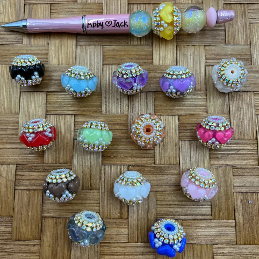 Assorted colorful fancy chunky beads decorated with AB rhinestone bands, pearl accents, and heart embellishments in black, blue, red, purple, pink, green, white, brown, and gray, displayed on a woven surface next to a decorated beadable pen.