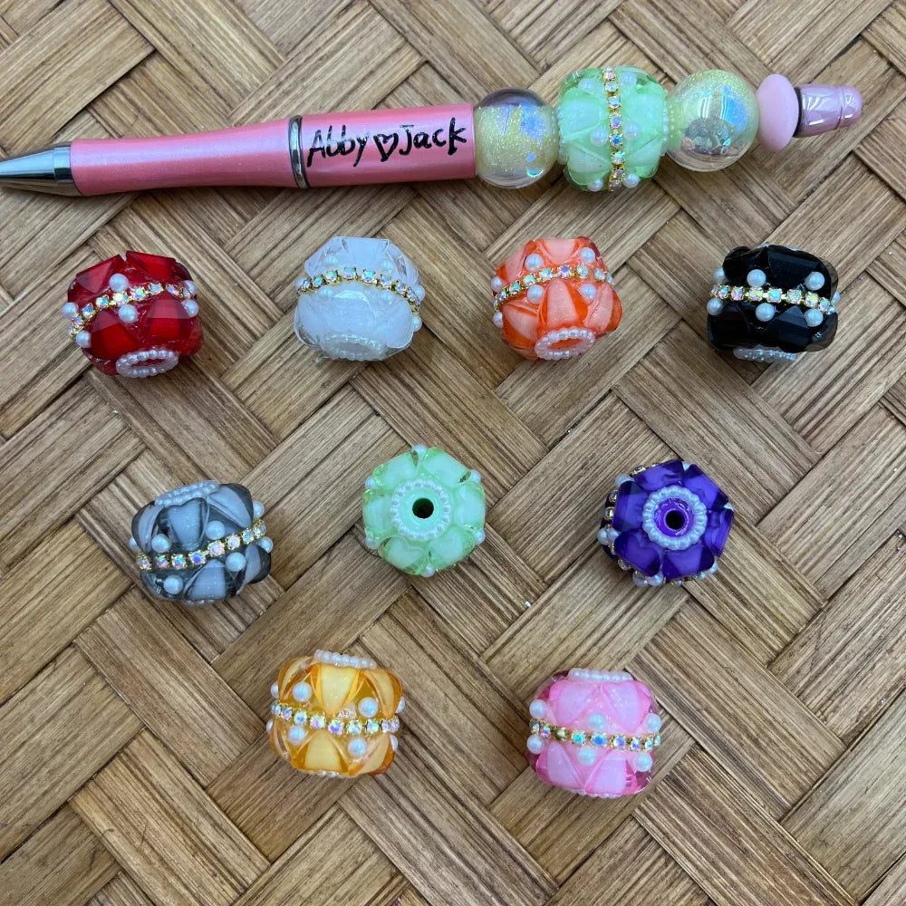 Assorted rhinestone and pearl fancy chunky beads in red, white, orange, black, gray, green, purple, yellow, and pink colors displayed on wicker surface next to a pink beadable pen labeled Abby Jack.