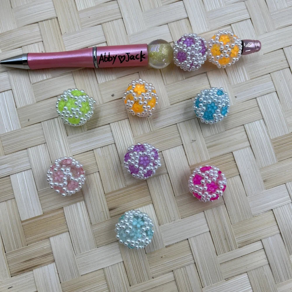 Colorful chunky round beads with white pearl studs and flower details in pastel pink, purple, teal, green, orange, and hot pink, displayed with a beadable pen on a woven surface.