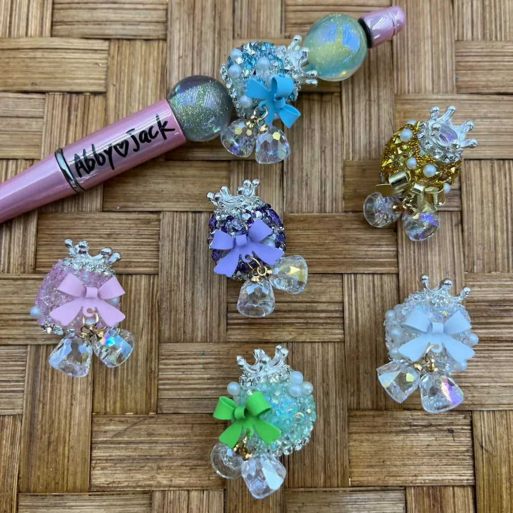 Assorted crown fancy beads with colorful bow charms and dangling crystal drops in pink, purple, blue, green, gold, and white, displayed on a wicker surface next to a decorated beadable pen labeled Abby Jack.