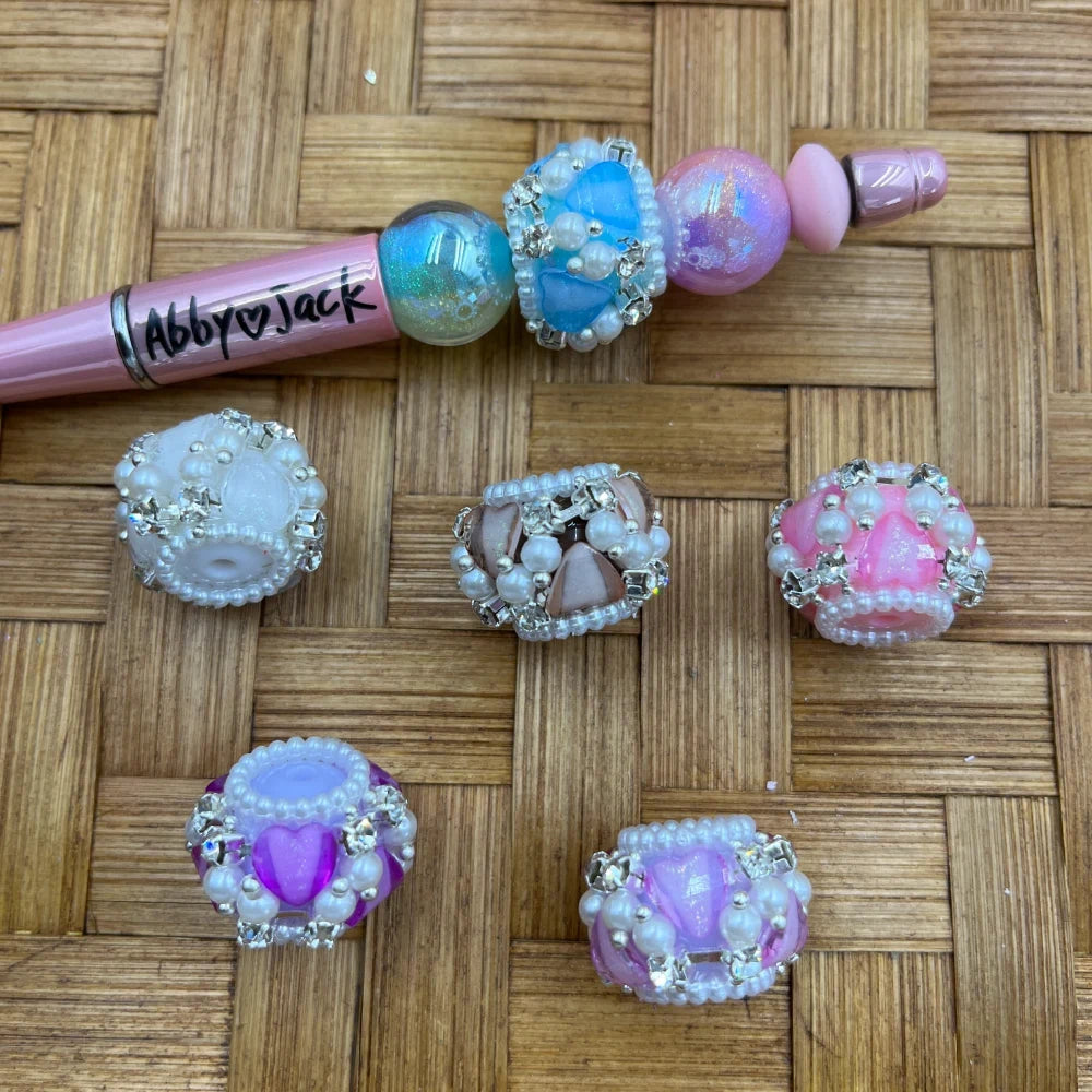 Assorted crown-shaped fancy beads decorated with rhinestones and pearl seed beads in pastel pink, purple, blue, white, and taupe colors, shown on a wicker surface alongside a pink beadable pen labeled Abby Jack.