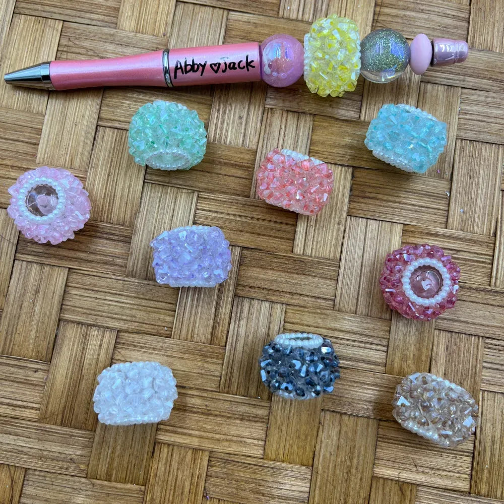 Assorted pastel crystal chunk rhinestone fancy beads in pink, green, lavender, teal, yellow, coral, white, gray and champagne displayed on a woven bamboo surface next to a pink beadable pen labeled Abby and Jack.