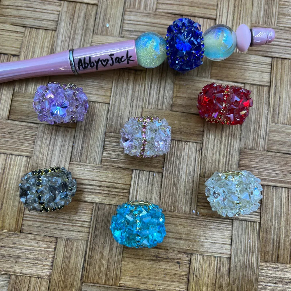 Assorted crystal cluster druzy chunky beads in purple, red, blue, teal, gray, clear, and white displayed on a woven surface next to a pink beadable pen labeled Abby Jack.