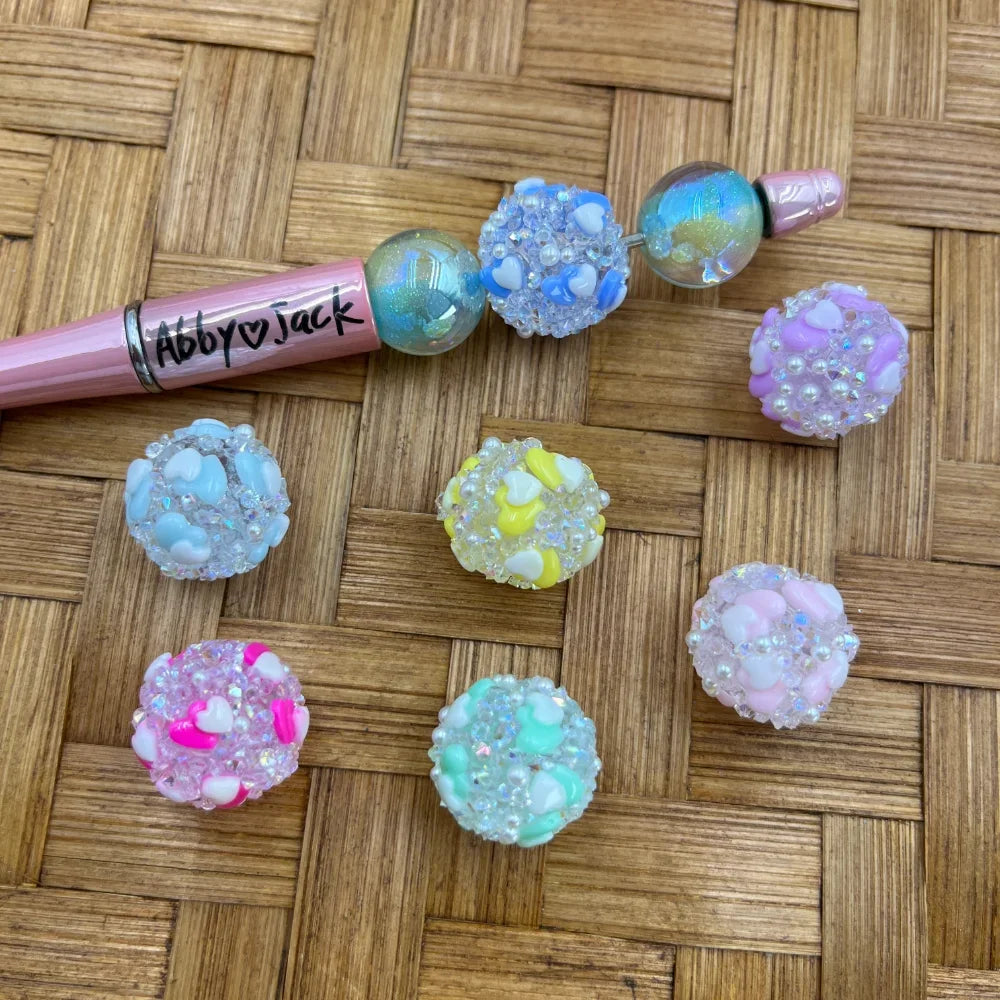 Assorted pastel crystal heart rhinestone fancy beads in blue, yellow, pink, mint, and lavender displayed on a woven surface next to a pink beadable pen branded Abby Jack, showing sparkly textured bead designs with heart embellishments.