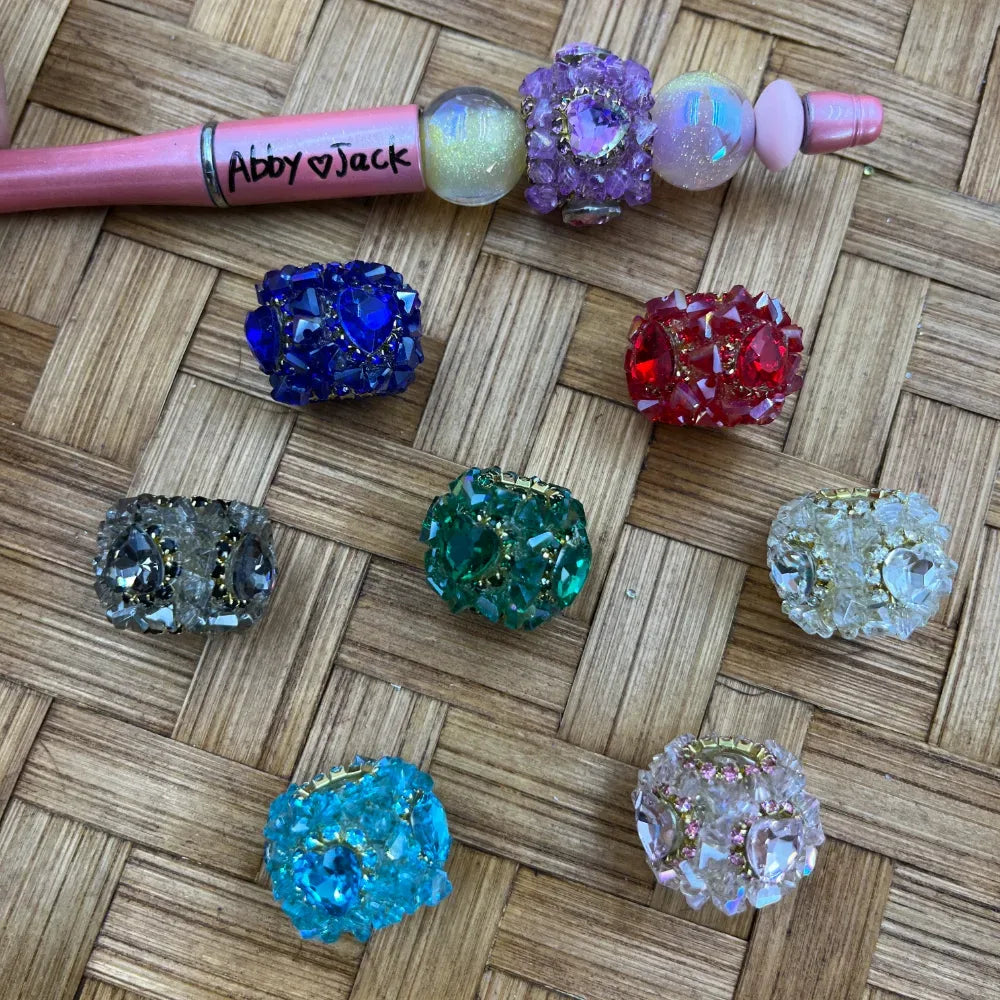 Assorted crystal rhinestone cluster fancy beads in blue, red, teal, green, gray, aqua, pink, and clear displayed on a woven surface next to a pink beadable pen labeled Abby and Jack