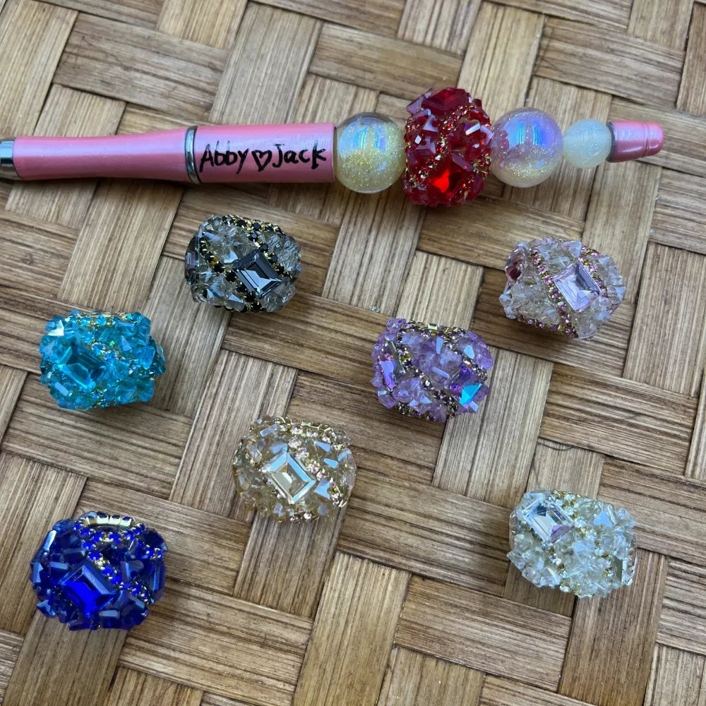 Assorted crystal rhinestone cluster fancy beads in blue, teal, purple, red, clear, lavender, and silver displayed on a wicker surface next to a pink Abby Jack beadable pen with iridescent globe beads.