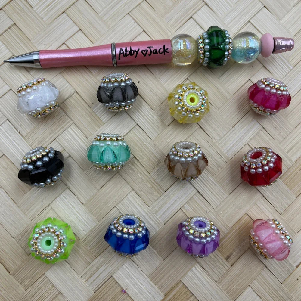 Assorted crystal rhinestone and pearl fancy beads in 12 colors including black, green, red, blue, purple, yellow, pink, and clear, displayed on woven bamboo mat with a decorated beadable pen labeled Abby and Jack.