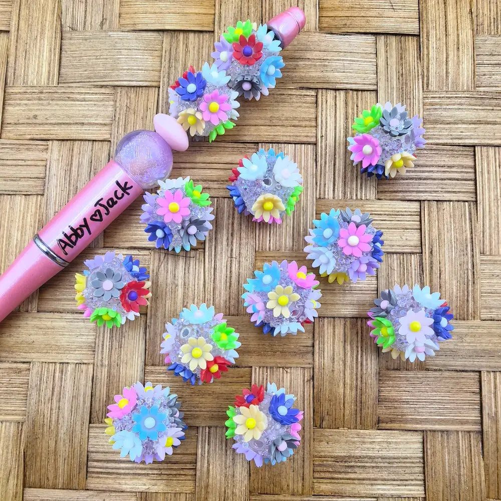 Colorful multicolor daisy flower cluster acrylic fancy beads displayed on woven bamboo surface next to a pink beadable pen labeled Abby Jack, showing pastel and bright floral bouquet bead designs.