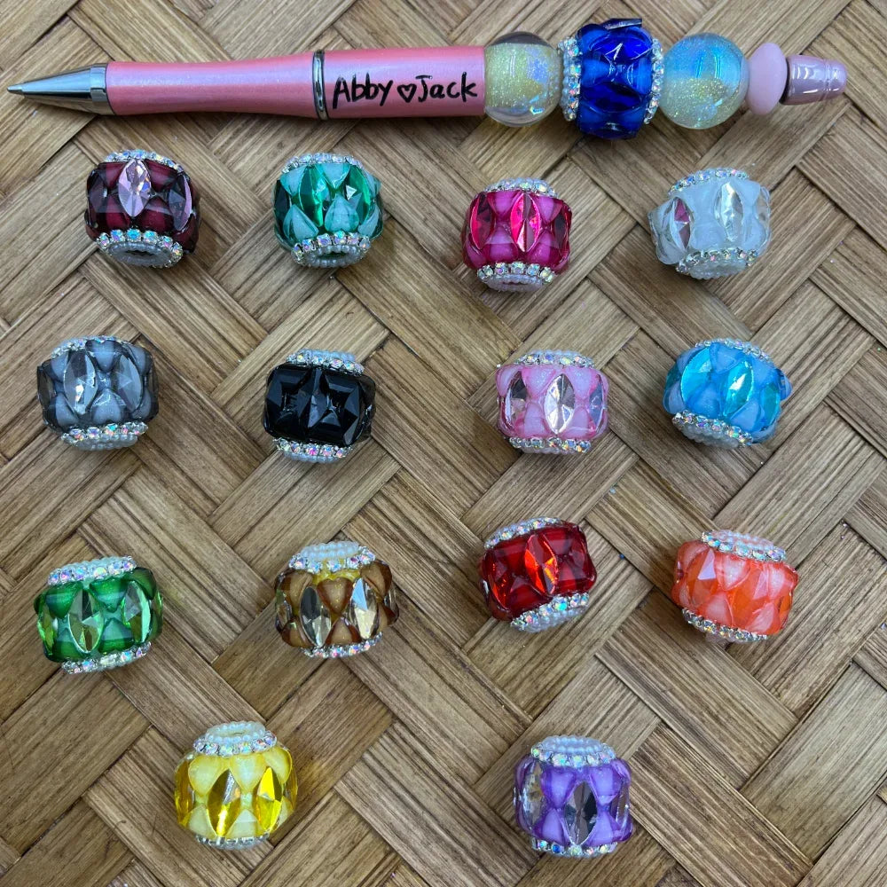 14 assorted faceted crystal rhinestone fancy beads in multiple colors including pink, green, blue, red, purple, yellow, orange, black, gray, and clear, displayed on a woven surface next to a decorative beaded pen labeled Abby and Jack.
