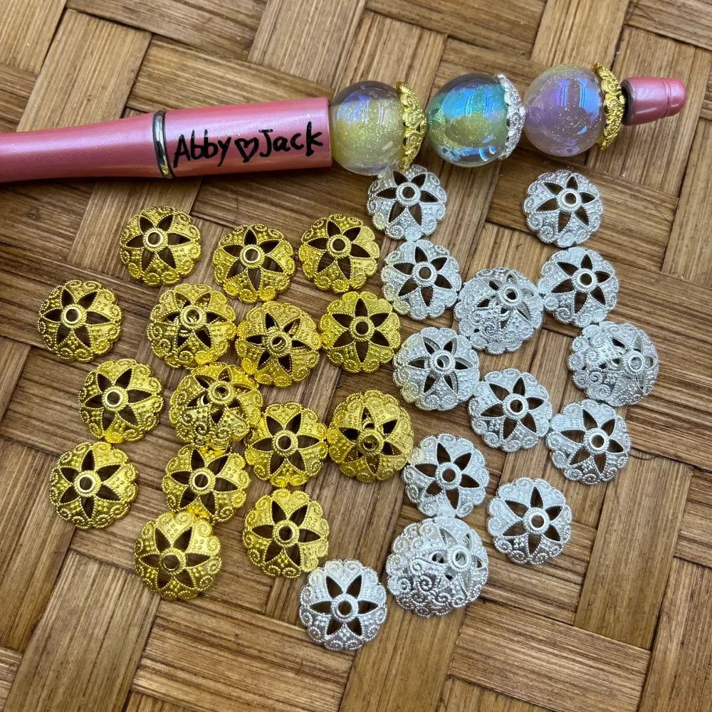 Gold and silver filigree flower bead caps displayed on a wicker surface next to a beadable pen decorated with iridescent globe beads and bead caps.