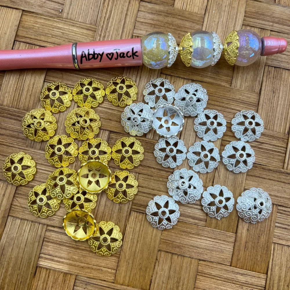 Gold and silver filigree flower metal bead caps displayed on woven surface next to a decorated beadable pen labeled Abby and Jack, showing intricate floral cutout pattern used for DIY jewelry making and beadable pens.